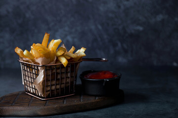 French fries in a metal basket with salt and ketchup on a stylish board on a dark background. Fried...