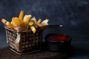 French fries in a metal basket with salt and ketchup on a stylish board on a dark background. Fried...