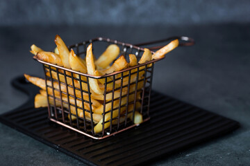 French fries in a metal basket with salt on a stylish board on a dark background. Fried potatoes....