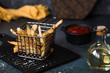 French fries in a metal basket with salt, ketchup and sunflower oil on a stylish board on a dark background. Fried potatoes. The concept of fast food and junk food.