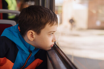 a cute boy in a bright sport jacket is riding alone on a tram and looks out the window. The boy's...