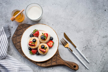 Cottage cheese pancakes, cheesecakes with fresh blueberries, strawberries, honey and mint leaves on a stylish wooden board on a gray concrete table. Natural products. Healthy and delicious breakfast.