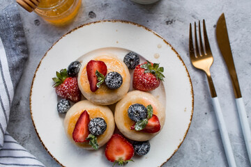 Cottage cheese pancakes, cheesecakes with fresh blueberries, strawberries, honey and mint leaves on a stylish wooden board on a gray concrete table. Natural products. Healthy and delicious breakfast.