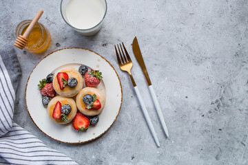 Cottage cheese pancakes, cheesecakes with fresh blueberries, strawberries, honey and mint leaves on a stylish wooden board on a gray concrete table. Natural products. Healthy and delicious breakfast.