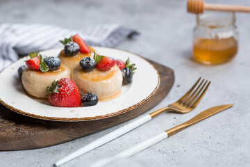 Cottage cheese pancakes, cheesecakes with fresh blueberries, strawberries, honey and mint leaves on a stylish wooden board on a gray concrete table. Natural products. Healthy and delicious breakfast.