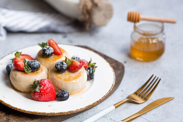 Cottage cheese pancakes, cheesecakes with fresh blueberries, strawberries, honey and mint leaves on a stylish wooden board on a gray concrete table. Natural products. Healthy and delicious breakfast.