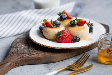 Cottage cheese pancakes, cheesecakes with fresh blueberries, strawberries, honey and mint leaves on a stylish wooden board on a gray concrete table. Natural products. Healthy and delicious breakfast.