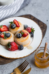 Cottage cheese pancakes, cheesecakes with fresh blueberries, strawberries, honey and mint leaves on a stylish wooden board on a gray concrete table. Natural products. Healthy and delicious breakfast.
