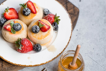 Cottage cheese pancakes, cheesecakes with fresh blueberries, strawberries, honey and mint leaves on a stylish wooden board on a gray concrete table. Natural products. Healthy and delicious breakfast.
