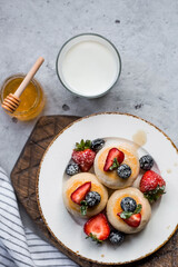 Cottage cheese pancakes, cheesecakes with fresh blueberries, strawberries, honey and mint leaves on a stylish wooden board on a gray concrete table. Natural products. Healthy and delicious breakfast.