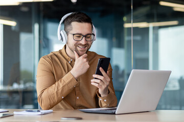 Young programmer working inside office with laptop, man in headphones smiling and listening to online music, audio podcast and audiobook, businessman in shirt at workplace holding phone