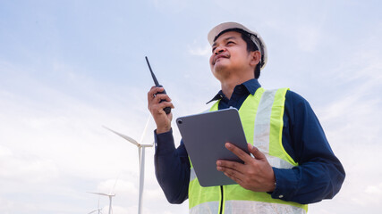 Asian man engineer wearing safety helmet, reflective vest holding tablet and walkie talkie with wind turbine propeller background.Energy and Environmental concept