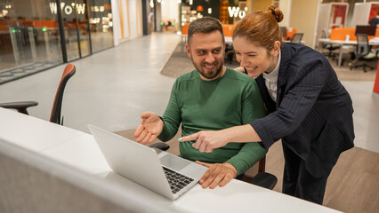 A red-haired Caucasian woman stands at the desktop of a bearded man. The boss approves the work of the subordinate. 