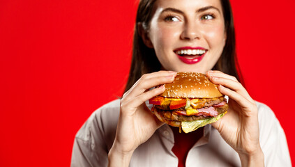 Happy young woman with big burger, bites hamburger with pleased smile, hungry face, stands isolated on red background