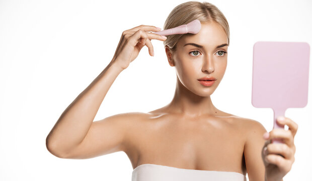 Young beauty woman after shower in towel, looks in mirror and applies makeup on clean glowing facial skin, using face brush to hide blemishes and imperfections with bb cream, white background