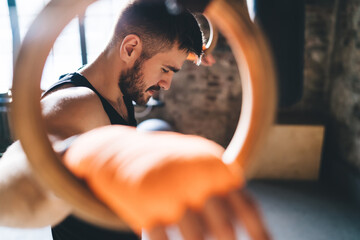 Focused young man taking break while standing with gymnastic rings in gym