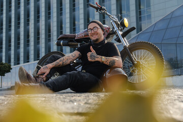 Shot of smiling stylish rocker with motorbike showing thumbs up sign outdoors.