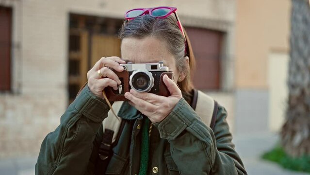 Mature hispanic woman with grey hair tourist wearing backpack taking pictures with vintage camera at street