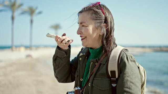 Mature hispanic woman with grey hair tourist wearing backpack speaking on the phone at seaside