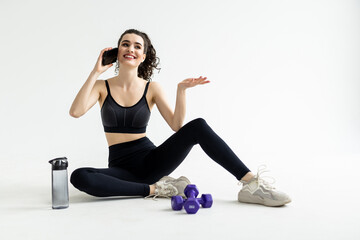 Morning exercise, a woman sits before performing exercises at home on the floor, talks on the phone happy and smiling, in a sports top and sweatpants. Healthy lifestyle, sports workout.