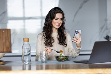 Beautiful sporty woman eating a bowl of salad while messaging with her mobile phone in the kitchen at home.