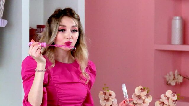 Pretty lady with neat make-up and hairstyle inflates soap bubbles in pink bathroom. Young woman in pink clothes stands in room with flowers