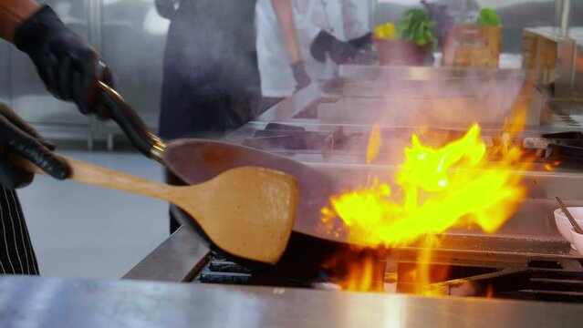 Closeup hands of chef man stir frying morning glory fire flame with burning pan in kitchen at restaurant, food and nutrition, cooking with movement slow motion, culinary and recipe.