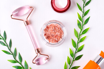 Skincare set with jade roller for facial massage, pink salt scrub, moisturizer and face serum in pump bottle on white background. Beauty treatment flat lay