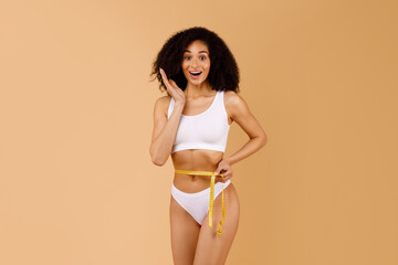 Body shaping concept. Shocked black lady measuring waist with tape, checking measures and being...