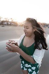 A young Hispanic woman is surprisingly looking at some seashells at the beach during sunset