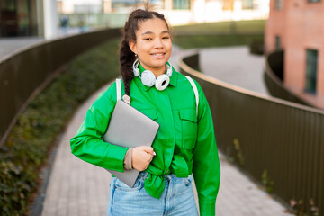 Happy mixed race lady walking in college campus with laptop and backpack, resting after classes outdoors