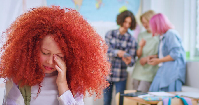 Crying girl with red curly hairstyle suffering from bullying by peers at school. Group of schoolchildren scoffing at their classmate on blur background.