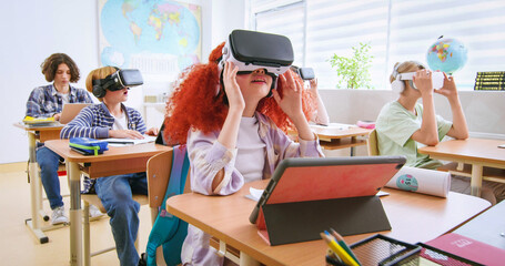 Interactive school concept. Kids in VR headsets sitting at desks and having virtual reality learning experience. Study technology. Kids in VR glasses in classroom. Tech education. - obrazy, fototapety, plakaty