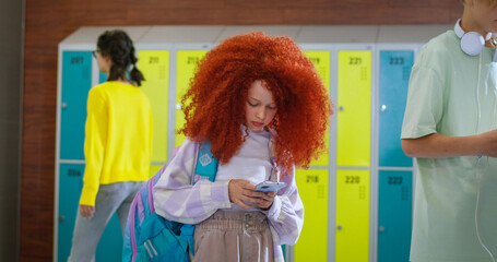 Redhead girl using modern smartphone in school hallway while other students walking around her. Female pupil with backpack standing alone during break and looking at camera.