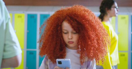 Portrait of cute Caucasian small girl with red curly hair standing at school corridor among people on a break, using smartphone and looking at camera. Little teen schoolgirl with a gadget.
