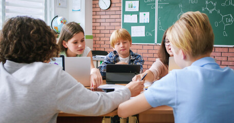 Enthusiastic female teacher using laptop for explaining chemical compounds to children during lesson. Attentive pupils looking at task on sheet of paper and making notes in workbook.