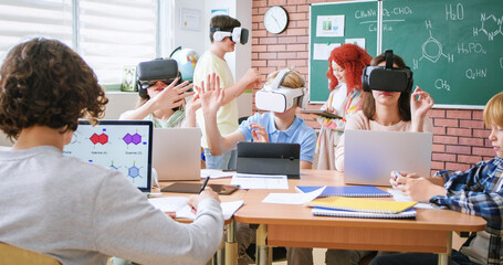 Elementary school age children sitting in VR glasses and learning to use virtual reality with help of teacher during chemical lesson. Concept of modern technology in education.