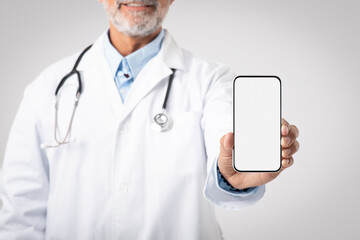 Smiling european senior man doctor in white coat with stethoscope shows smartphone with blank screen