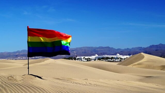 Bandiera arcobaleno sventolando con orgoglio all'aperto tra dune le di sabbia, la montagna sullo  sfondo e il cielo azzurro di Maspalomas 