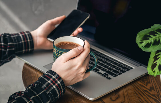 A cup of cocoa and a smartphone in the hands of a man in front of a laptop.