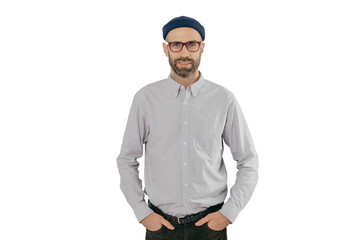 Indoor shot of satisfied Caucasian man in spectacles with bristle, keeps hands in pockets, dressed in formal stylish clothes, has gentle smile, happy to be promoted, isolated over white background.