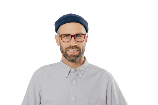 Horizontal view of pleased smiling man wears glasses, hat and shirt, being satisfied with good news, models against white background. Unshaven hipster rejoices new purchase. Fashion concept.