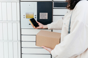 A woman with a phone near the self-service mail terminal. Parcel delivery machine. Person holding a cardboard box. Mail delivery and post service, online shopping, e commerce concept