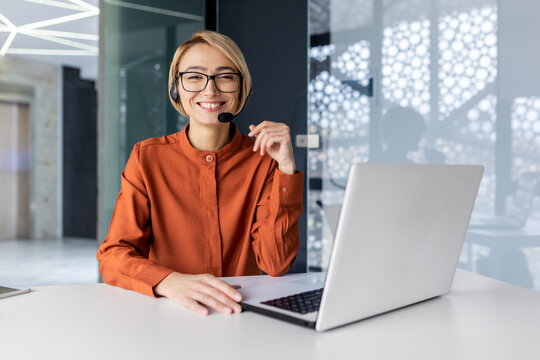 Portrait of successful online customer support office worker, satisfied business woman at workplace with video call headset and laptop smiling and looking at camera.