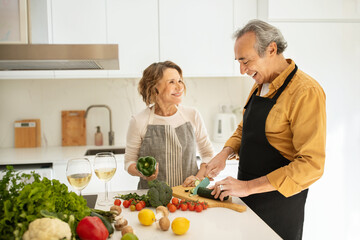 Happy elderly spouses cooking together and talking, woman and man preparing vegetable salad, kitchen interior