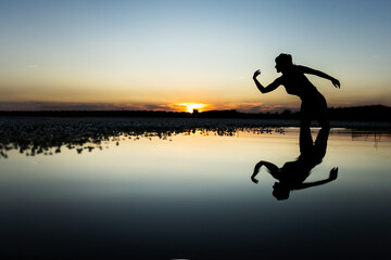 woman in the lake making figures and dancing with her body in the water, at sunset, with cold and warm colors in the sky, reflections in the water.