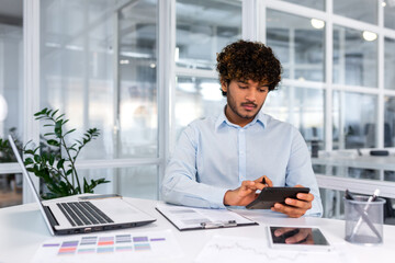 Serious thinking hispanic businessman working with documents and invoices inside office, man behind...