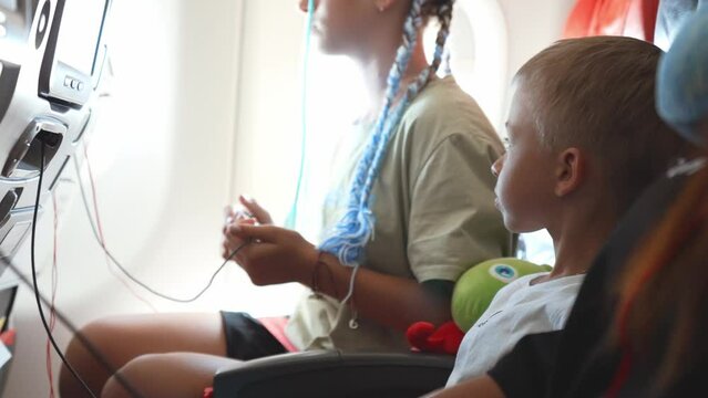 Kid boy playing video game with gamepad in hands looks at screen in airplane during long flight. Teen sister girl sits near looks at window on clouds. Family flight with children on vacation concept.