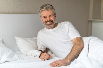 Happy handsome middle aged man sitting in bed, smiling