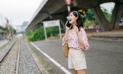 Asian young woman traveler with weaving basket using a mobile phone beside railway train station in Bangkok. Journey trip lifestyle, world travel explorer or Asia summer tourism concept.